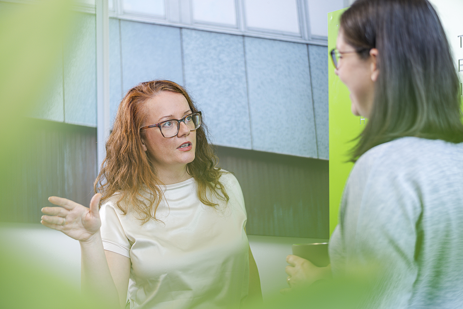 Close-up of two colleagues talking in a brightly lit office area with modern glass architecture in the background.