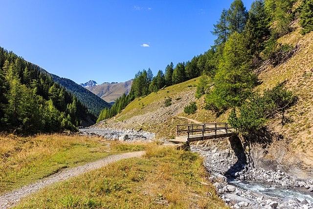 In a mountainous daytime landscape, a small wooden bridge spans the rocky bed of a swiftly flowing stream. It can carry only a small amount of foot traffic.